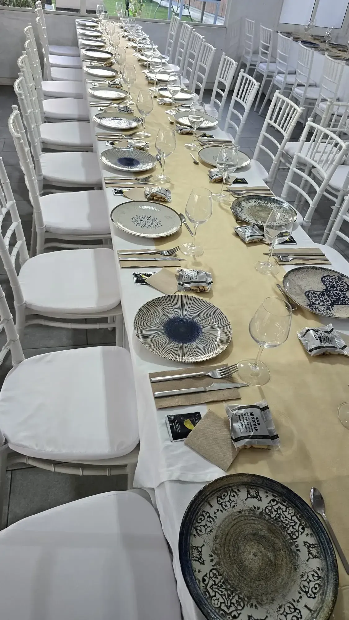 Mesa larga de banquete puesta con platos estampados, cristalería, cubiertos y sillas blancas en una habitación luminosa.
