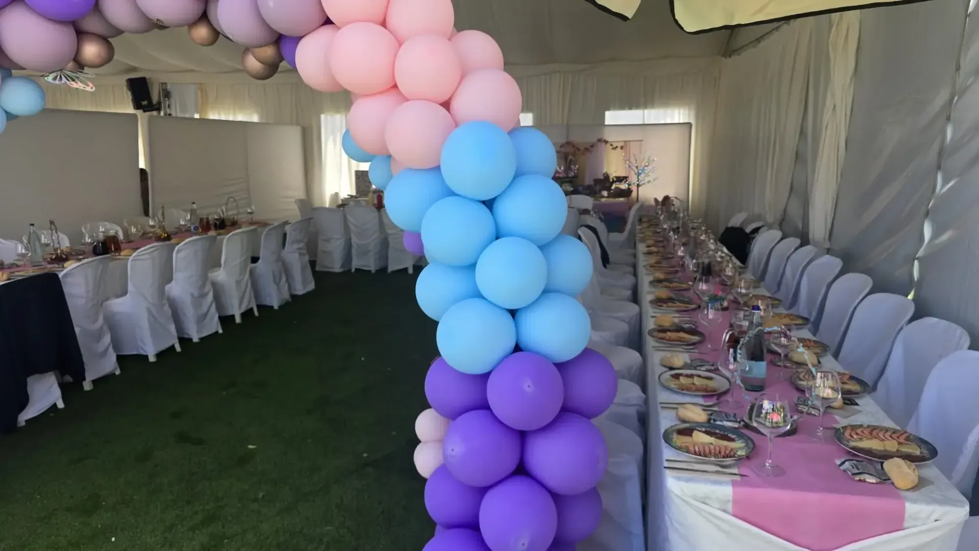 Un arco de globos en tonos rosa, azul y morado decora una carpa de fiesta con largas mesas dispuestas para los invitados.