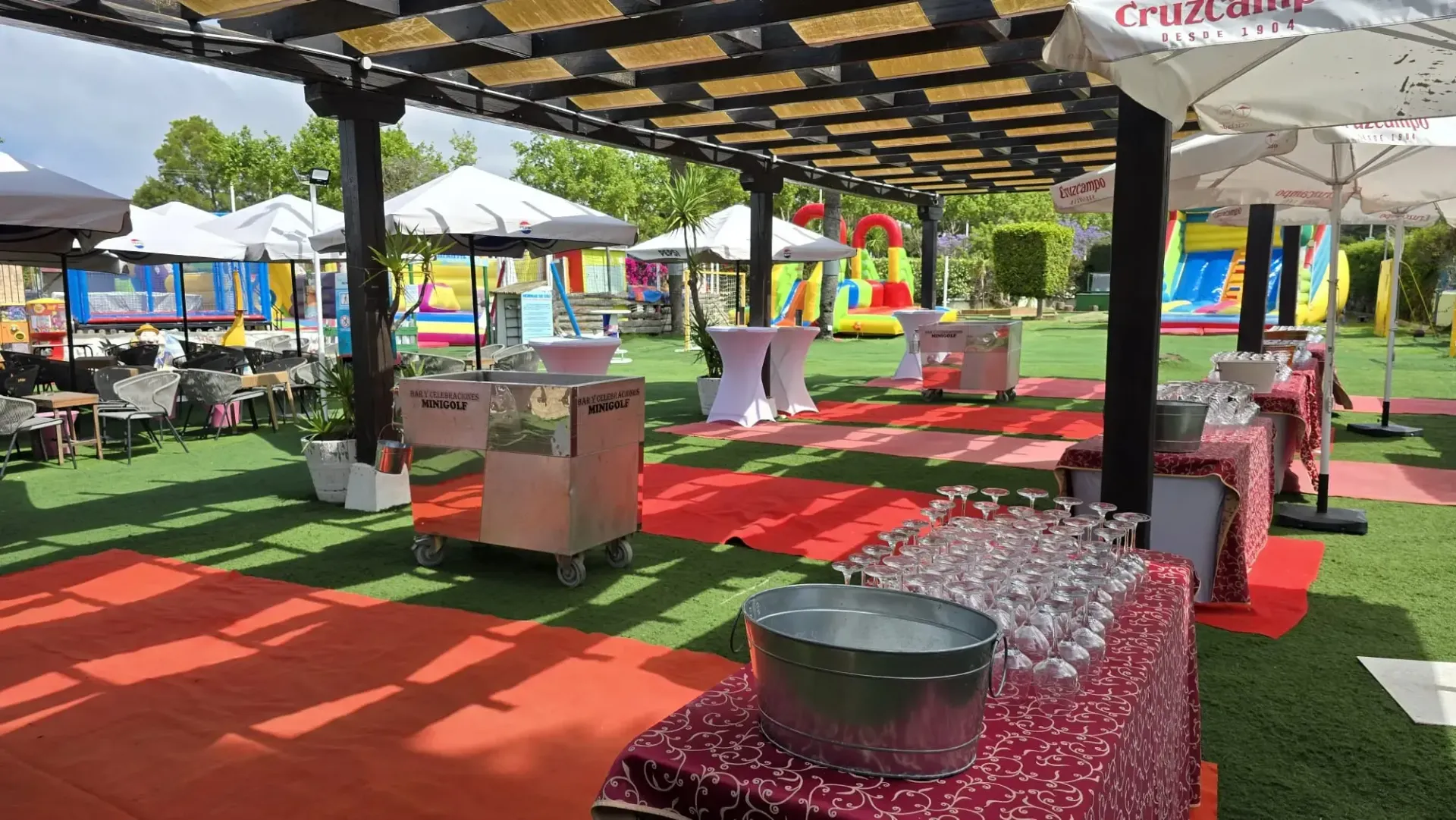 Zona de estar al aire libre con alfombra roja, bancos estampados, sombrillas y mesas bajo una pérgola.