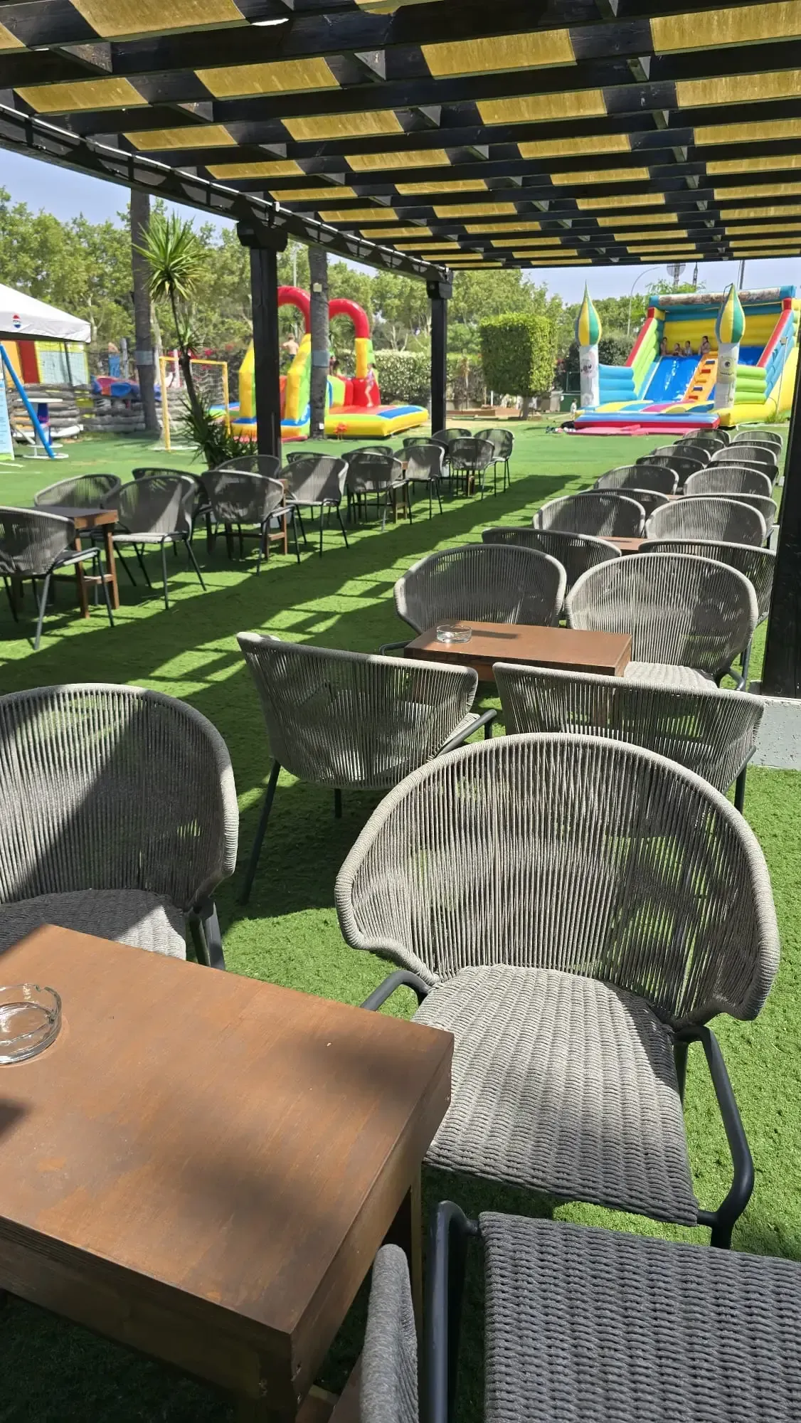 Zona de estar al aire libre en el patio con mesas y sillas bajo un toldo a rayas junto a un colorido tobogán inflable.