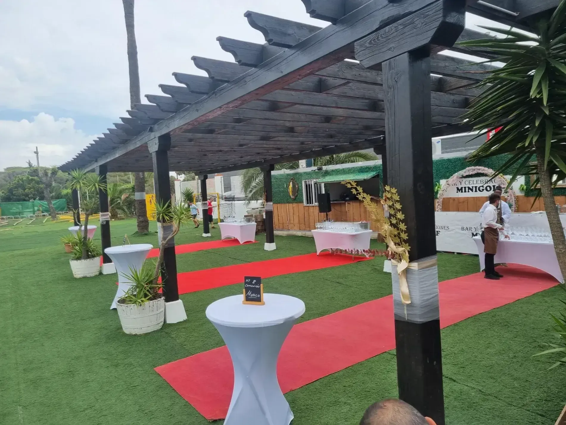 Un hombre está de pie bajo una pérgola con mesas y una alfombra roja.