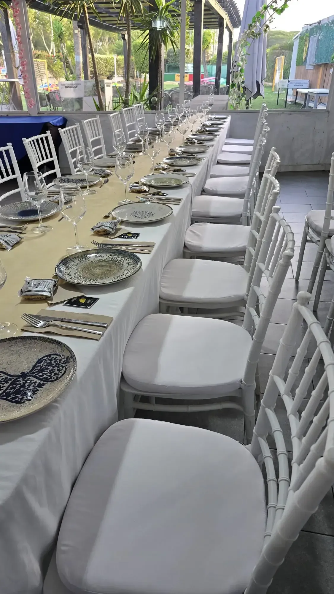 Conjunto de mesa de comedor larga para exteriores con sillas blancas y platos estampados bajo un patio cubierto.