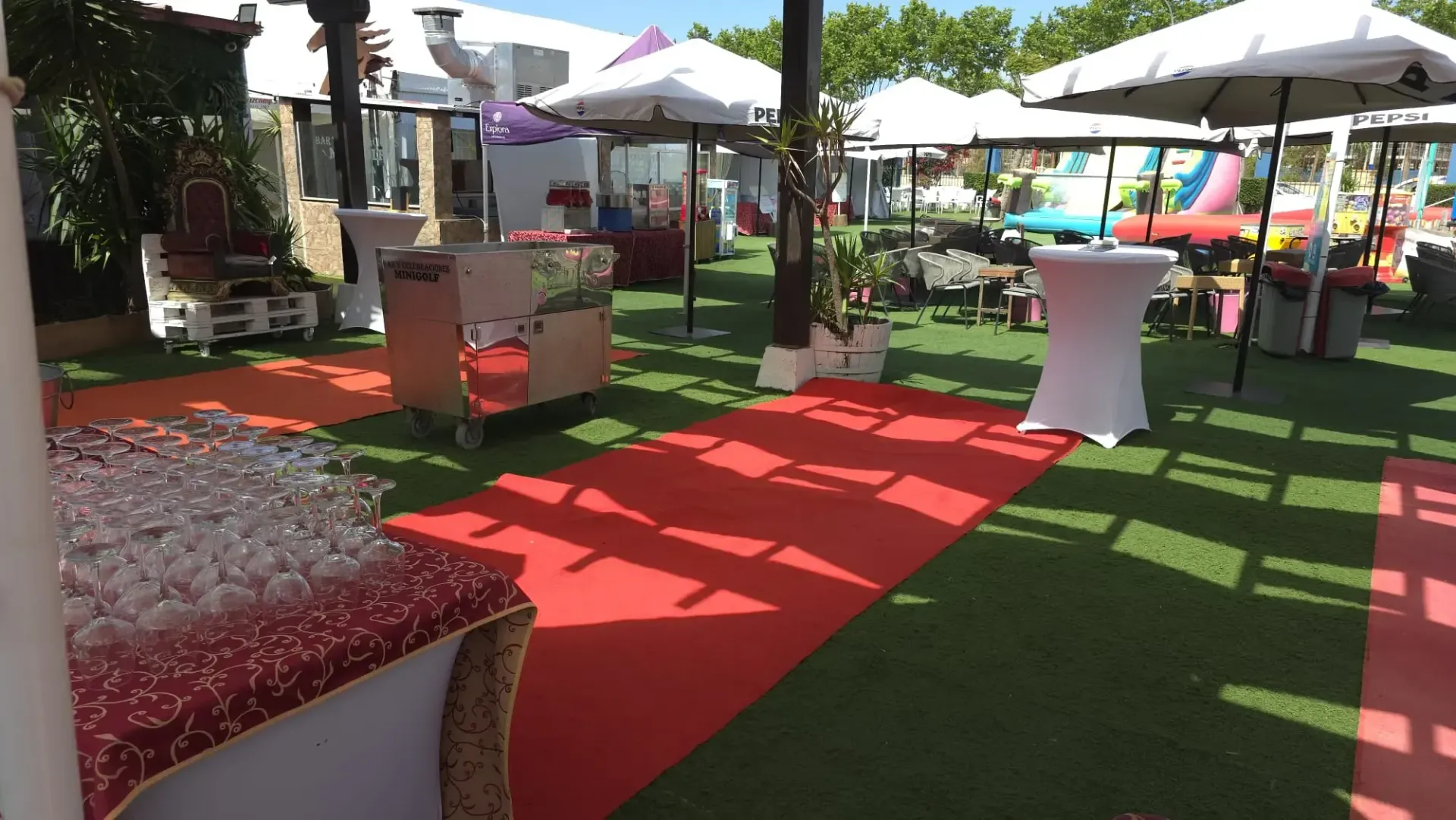 Salón al aire libre con alfombra roja, mesas de cóctel blancas, sombrillas y asientos sobre el césped en un jardín soleado.