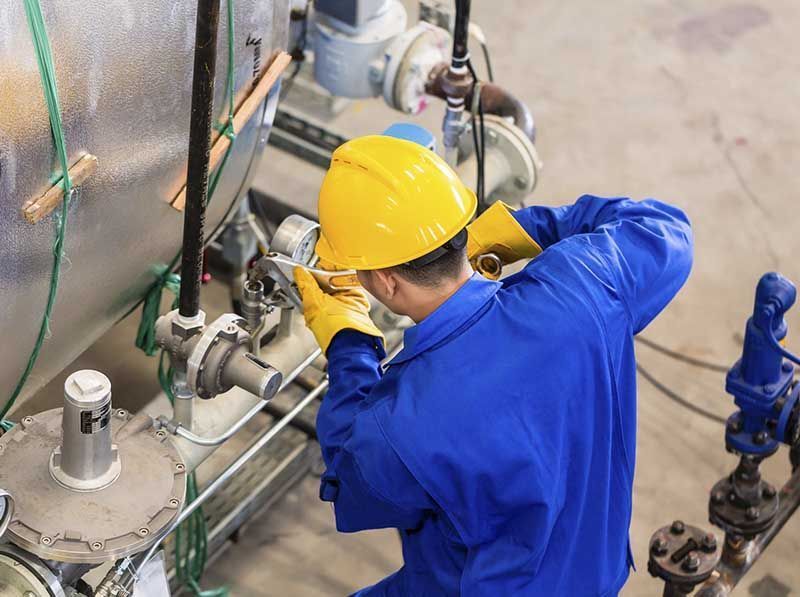 Un trabajador con mono azul y casco amarillo inspecciona tuberías y válvulas industriales en una fábrica.