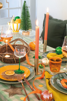 Table dressée avec de la vaisselle orange et verte, des bougies orange allumées et de la décoration de Noël disposée entre les couverts.
