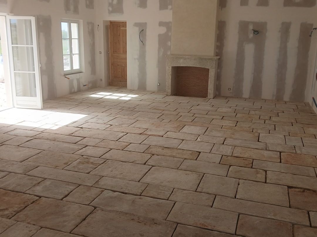 Pièce vide et inachevée avec sol en pierre, murs apparents, cheminée et lumière vive provenant d'une fenêtre et d'une porte ouverte.