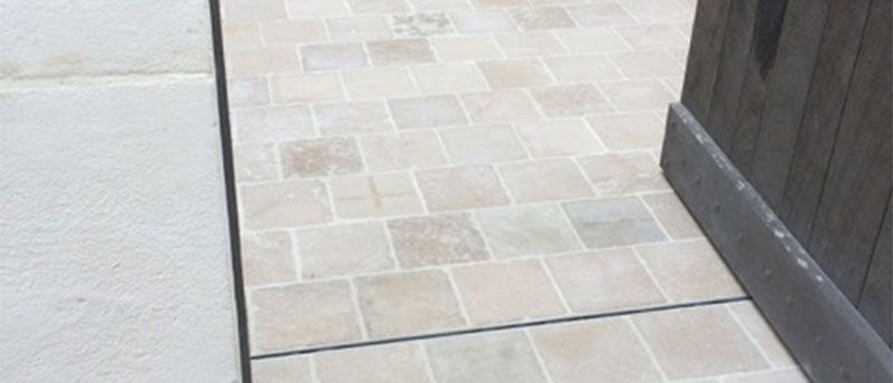 Terrasse carrelée beige le long d'un mur clair, avec un encadrement de porte en bois visible sur la droite.