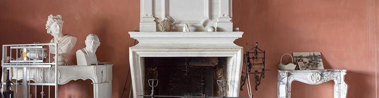 Une cheminée blanche, un mur rouge et une table en marbre. Divers objets décoratifs ornent la cheminée, la table et les abords du foyer.