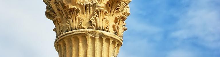 Gros plan sur une colonne corinthienne en pierre sculptée, beige, se détachant sur un ciel bleu et blanc.