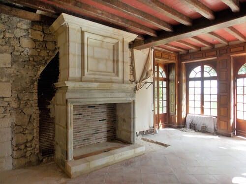 Intérieur de la pièce en rénovation : cheminée en pierre, briques apparentes, lambris, fenêtres cintrées, poutres apparentes au plafond.