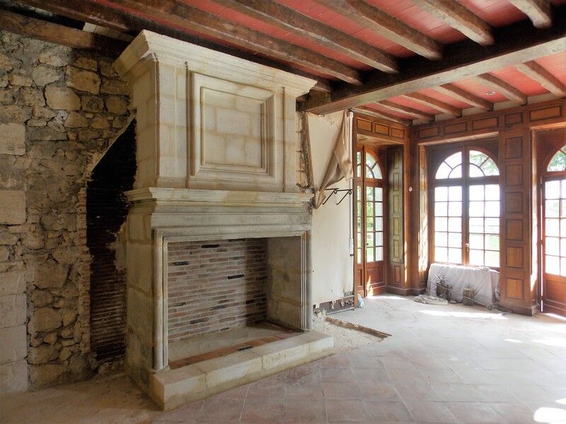 Pièce rustique avec cheminée en pierre, poutres apparentes et grandes fenêtres laissant entrer la lumière du jour