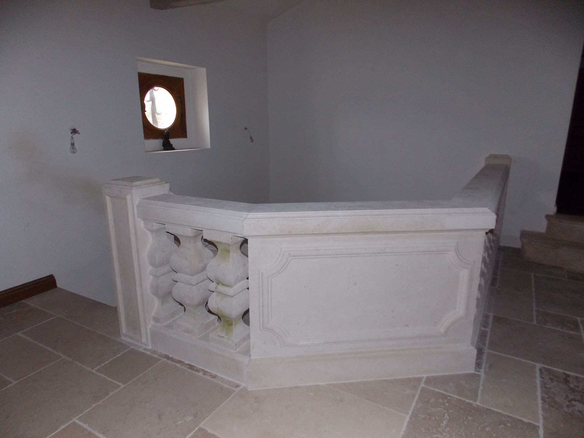 Intérieur vide avec un bar ou un comptoir en pierre blanche et un sol carrelé, à côté d'un escalier et d'une petite fenêtre.