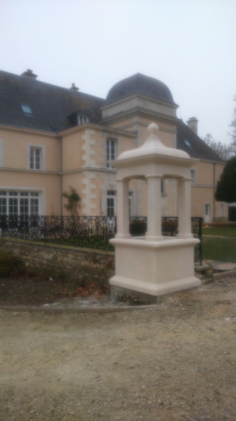 Petit monument en pierre blanche devant un château beige au toit sombre et à la clôture en fer forgé.
