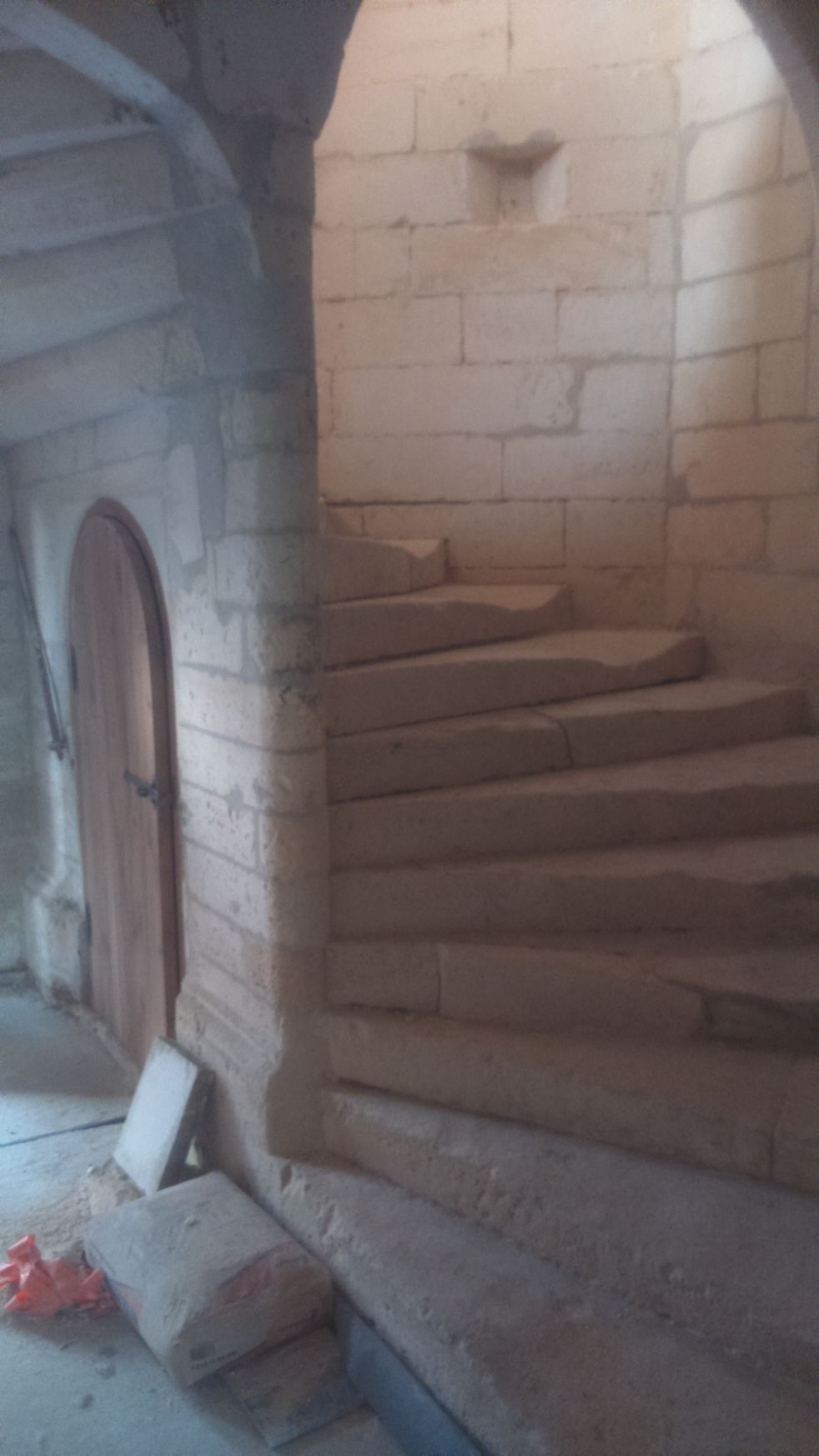 Escalier en colimaçon de pierre à l'intérieur d'un couloir en maçonnerie inachevée, avec une porte cintrée et des gravats au sol.
