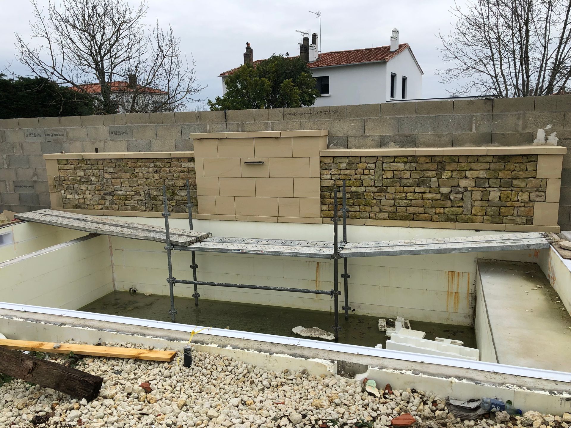Piscine en construction, partiellement vidée, dans une cour arrière, avec un mur de pierre et une maison en arrière-plan.