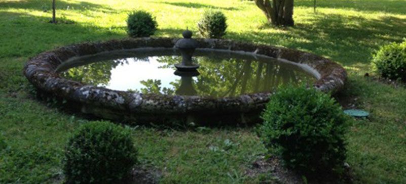 Fontaine circulaire en pierre avec un ornement central, entourée d'herbe verte et d'arbustes.