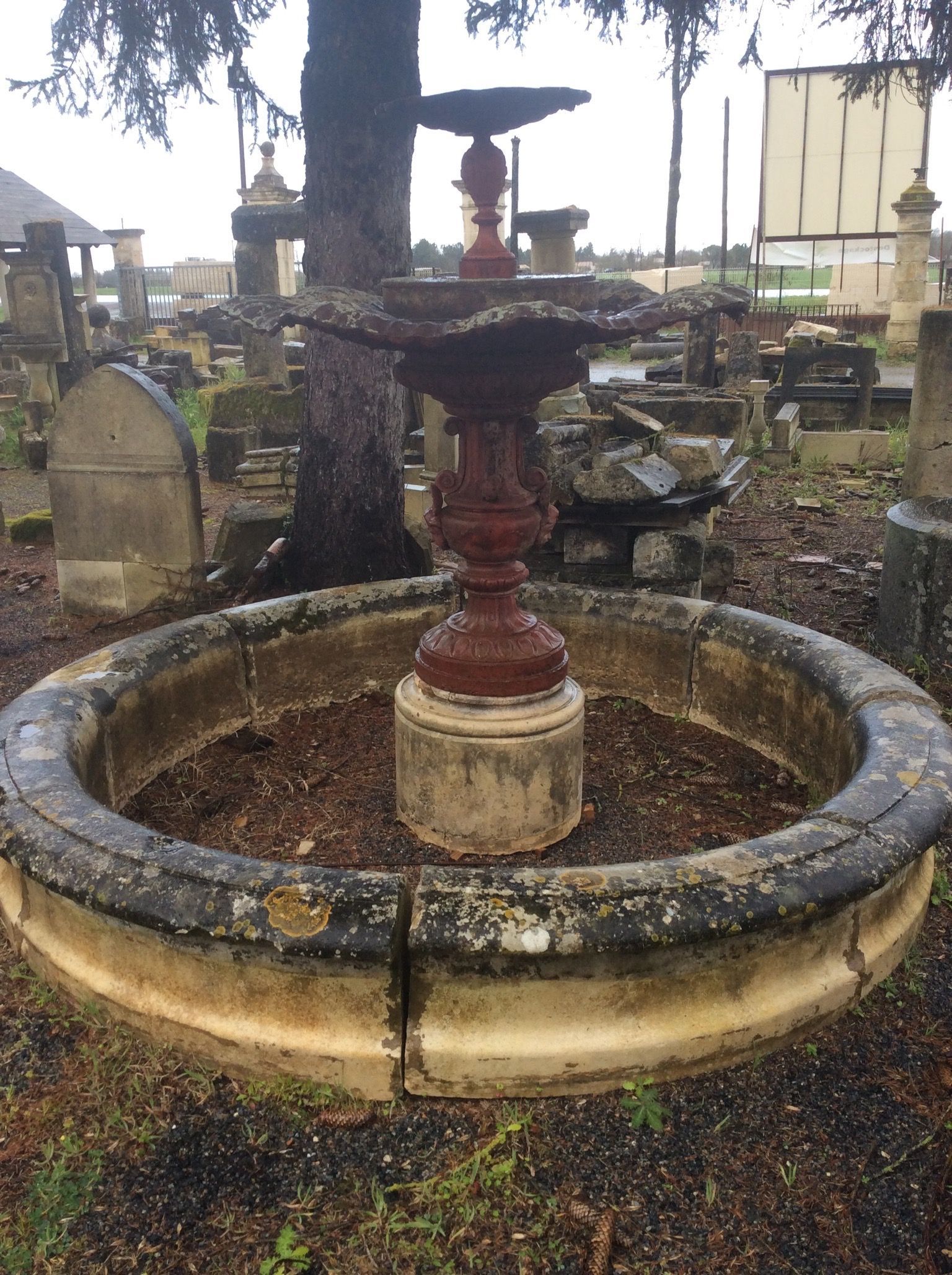 Vieille fontaine en pierre dans un cimetière, avec un piédestal rouge et un bassin circulaire parmi les pierres tombales.