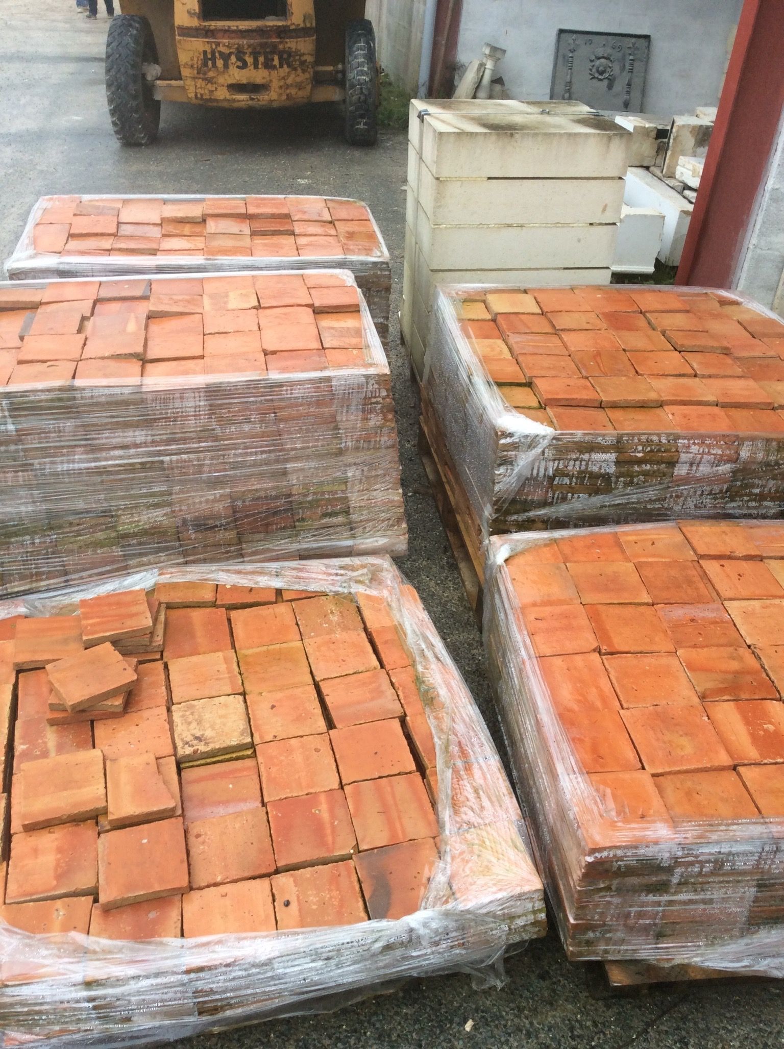 Des piles de briques orange emballées dans du plastique sont entreposées à côté d'un camion dans une cour extérieure.