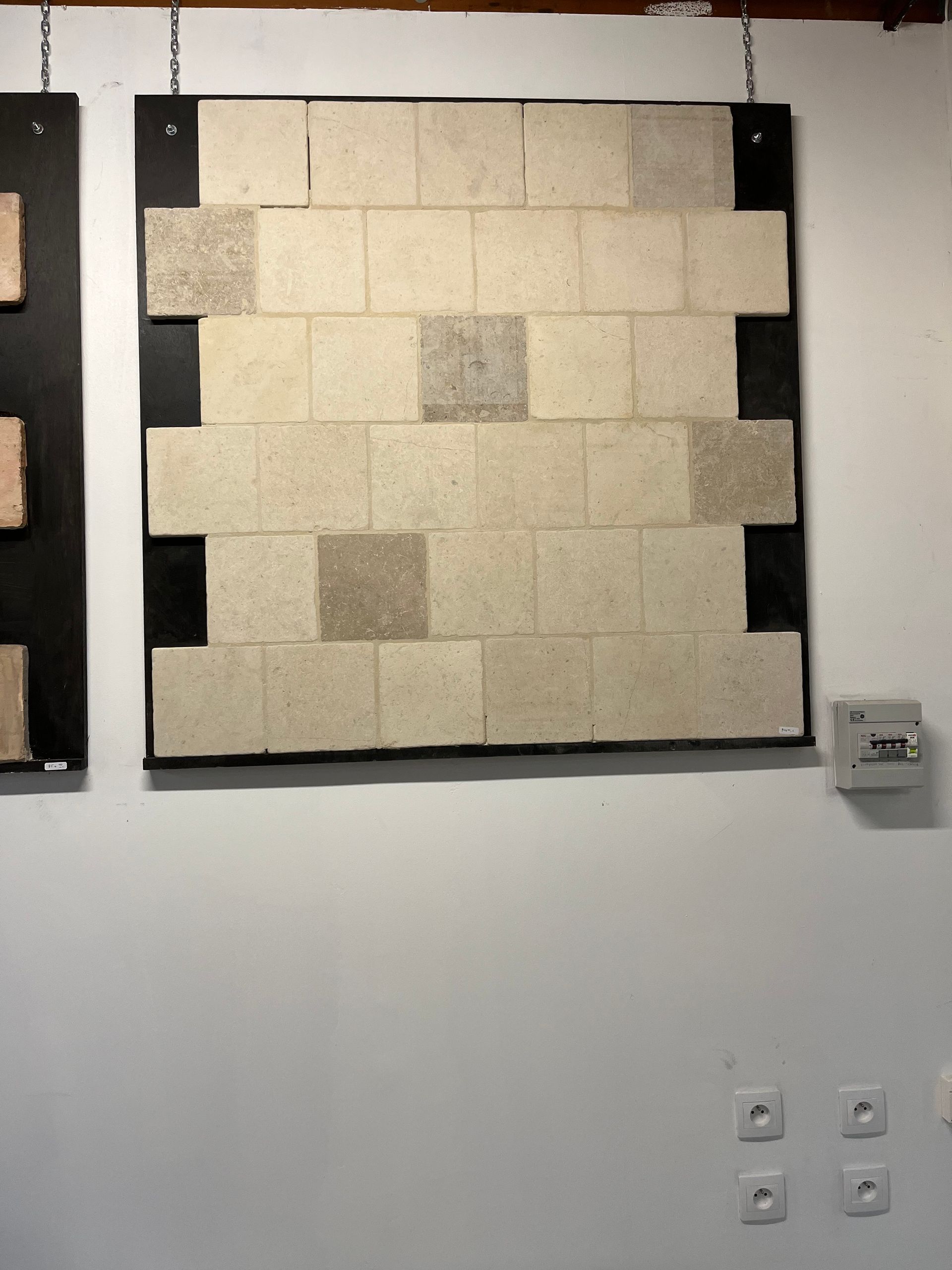 Composition murale de carreaux beiges encadrés de noir, fixée sur un mur de galerie blanc.