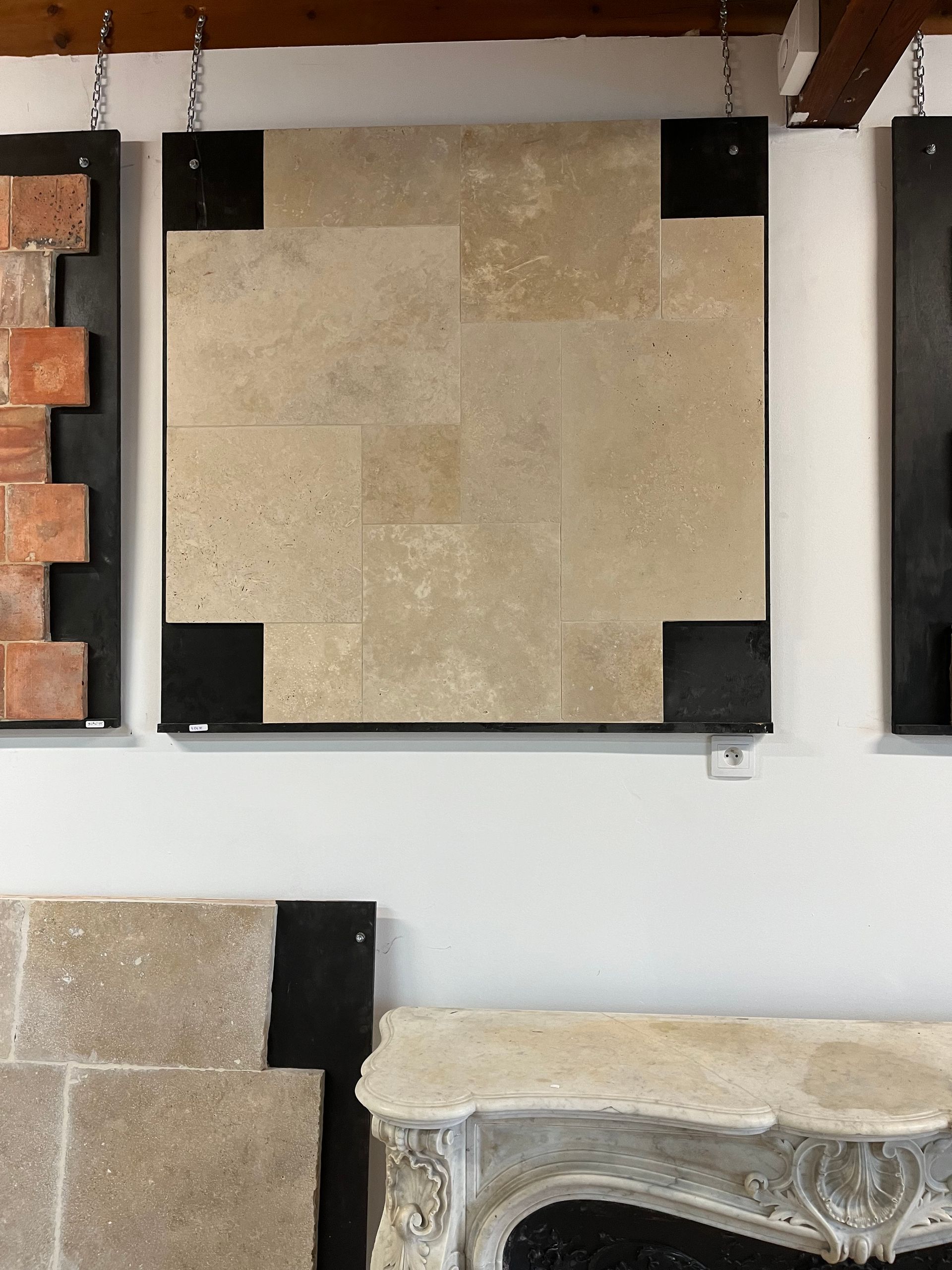 Présentation murale d'échantillons de carreaux de pierre beige avec supports d'angle noirs dans une salle d'exposition.