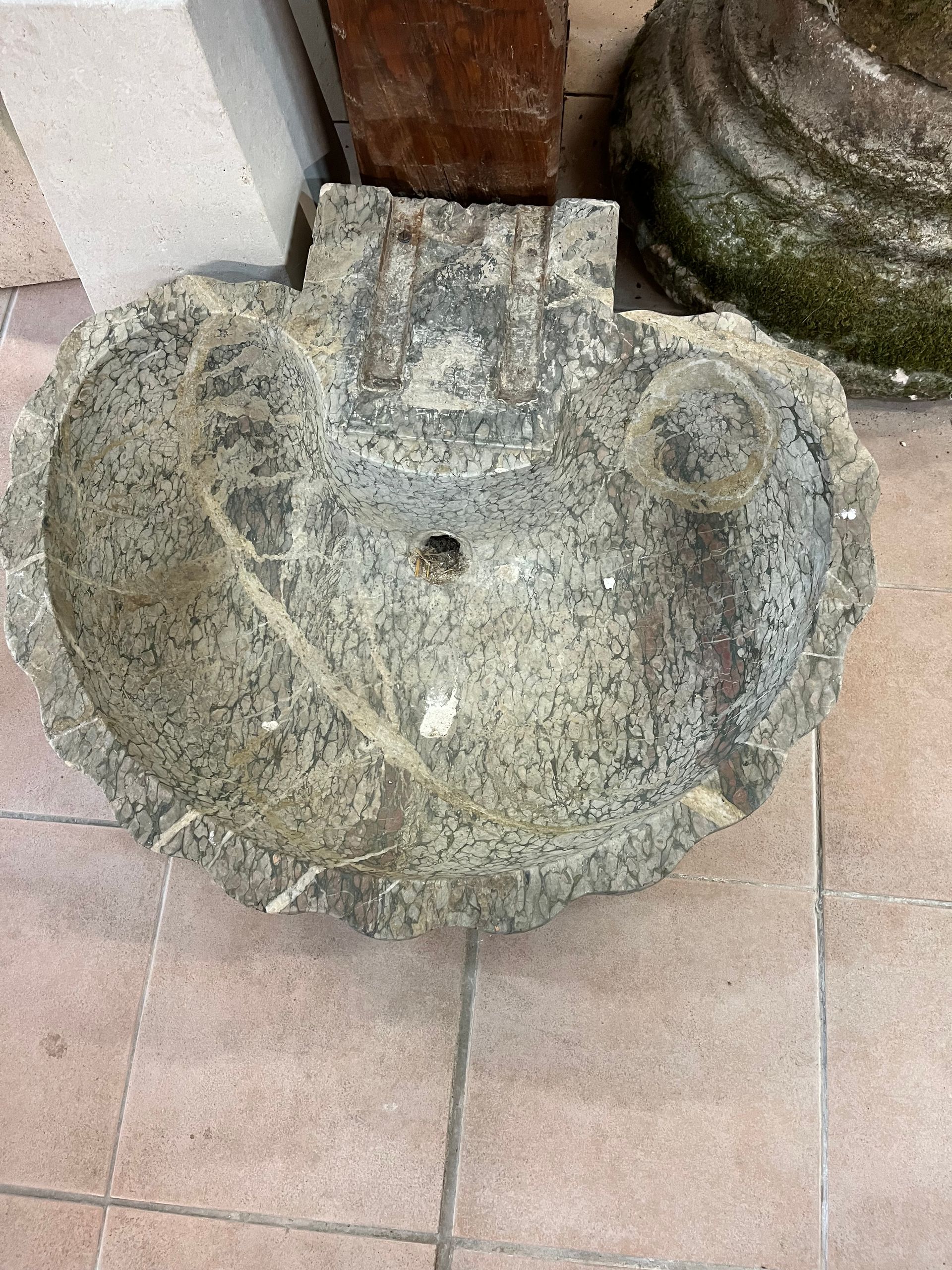 Lavabo ou vasque en pierre patinée sur un sol carrelé, à côté d'un mur et d'une porte en bois.