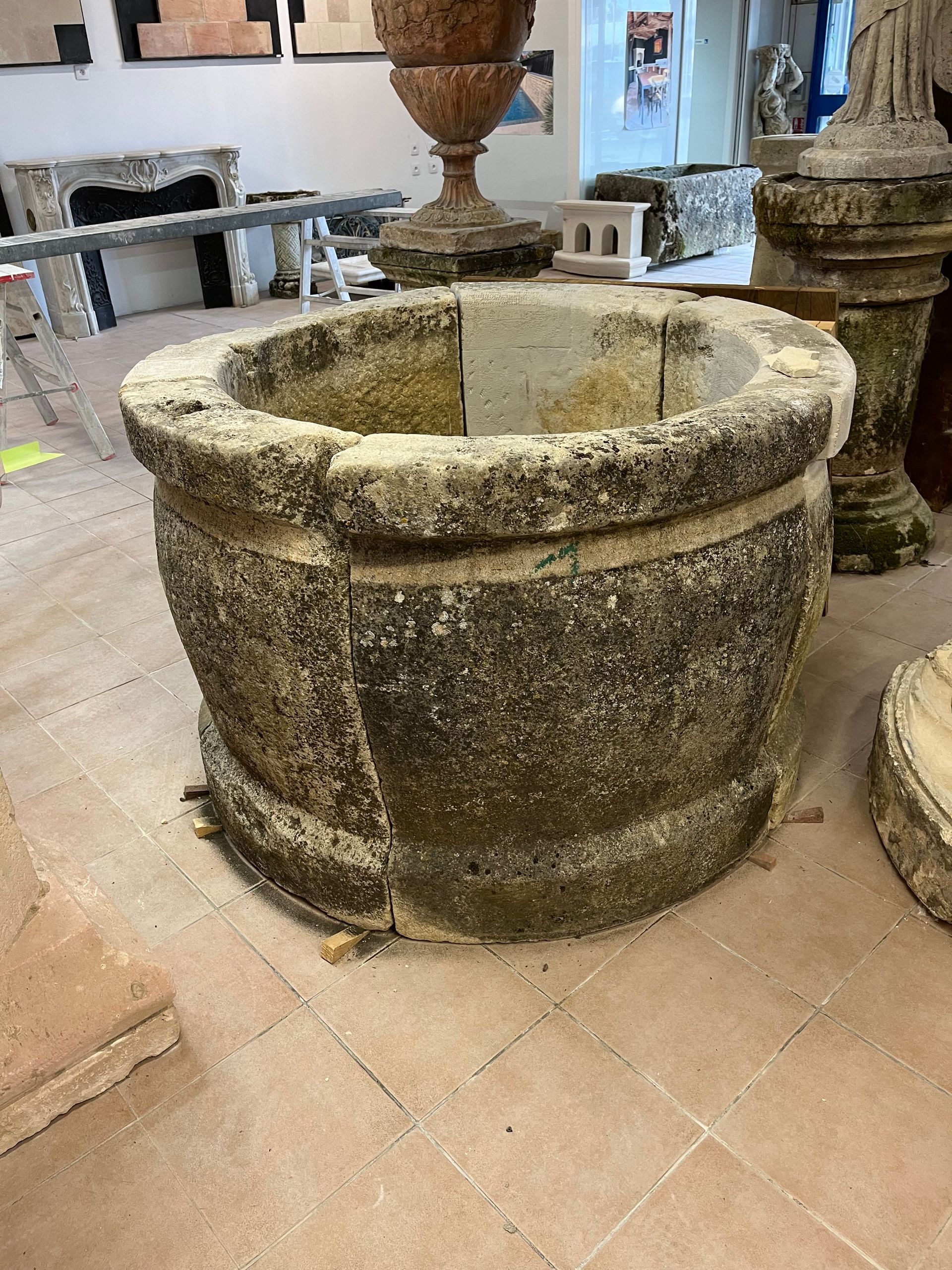 Grande fontaine ou jardinière en pierre patinée dans une cour intérieure carrelée, avec d'autres éléments en pierre à proximité.