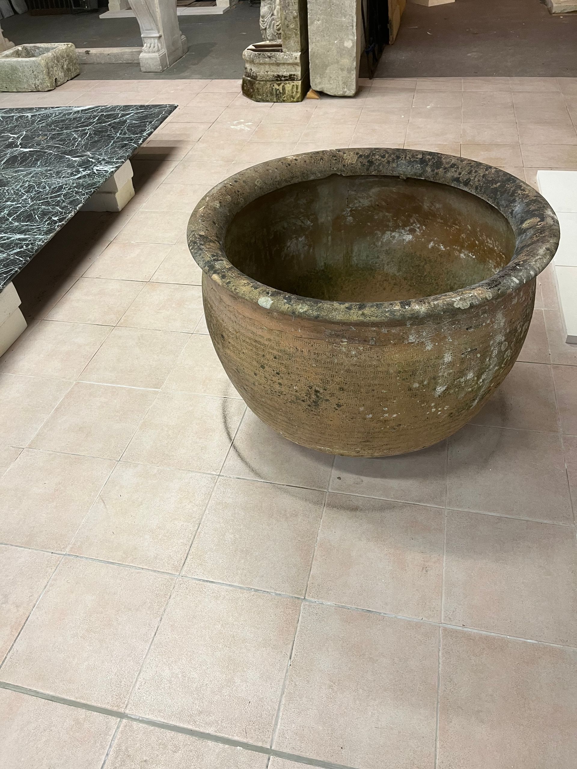 Pot de fleurs en pierre patinée sur une terrasse carrelée, à la surface brun mousseux et à l'intérieur vide.