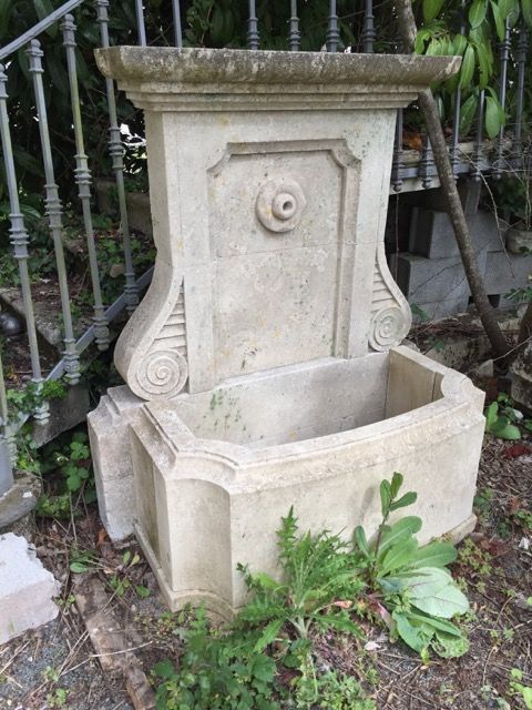 Ancienne fontaine de jardin en pierre avec un bassin, adossée à une clôture en fer forgé et entourée de plantes