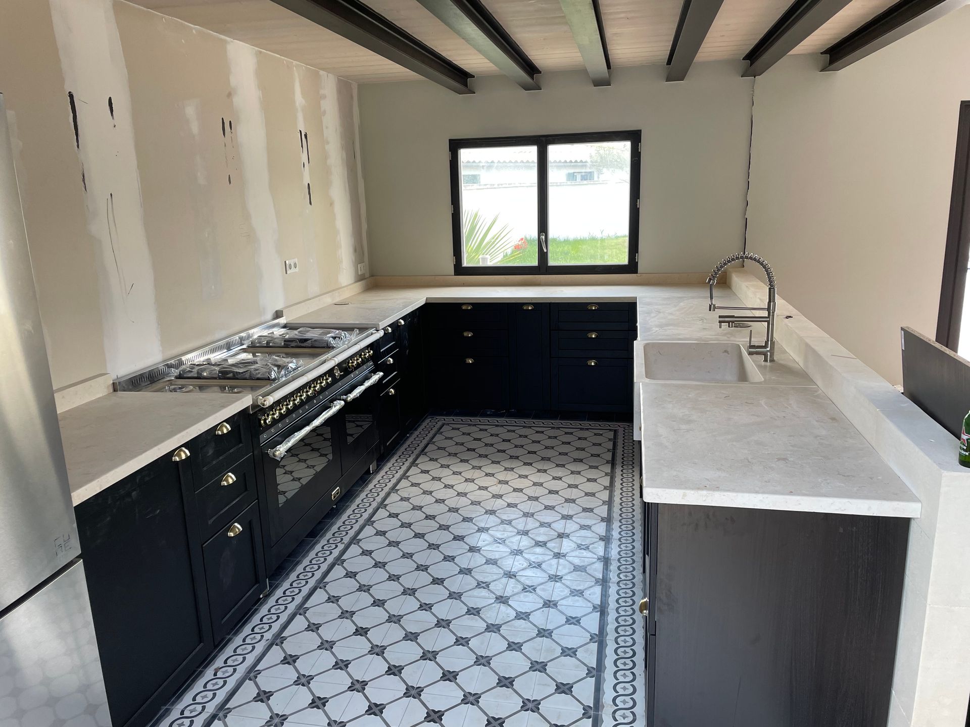 Cuisine étroite avec placards noirs, plans de travail blancs, sol carrelé à motifs et une fenêtre au fond.