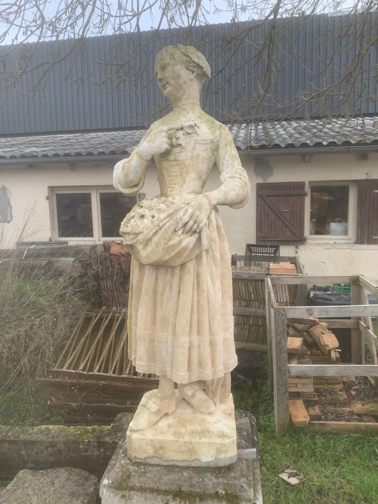 Statue en pierre patinée représentant une femme tenant des fleurs, dressée à l'extérieur, à côté d'une maison rustique.