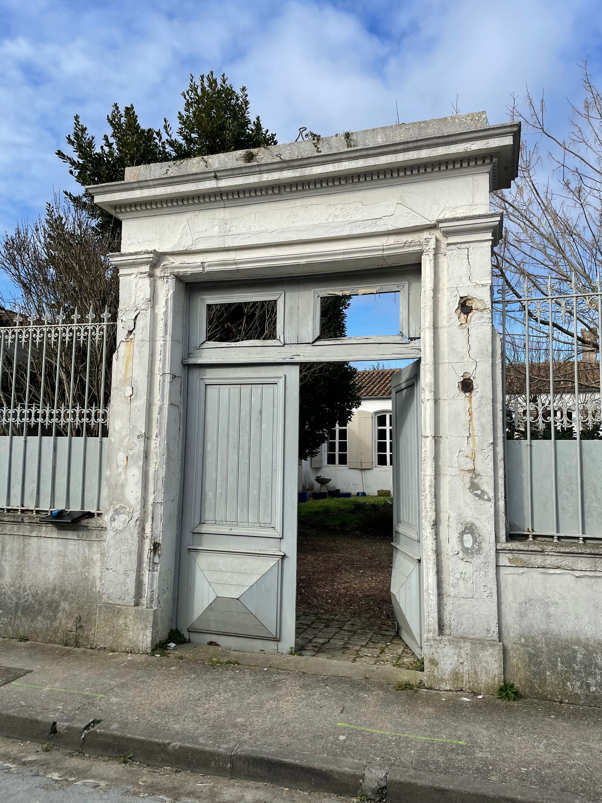 Un vieux portail en pierre avec une porte blanche entrouverte, donnant sur une cour intérieure derrière une clôture métallique.