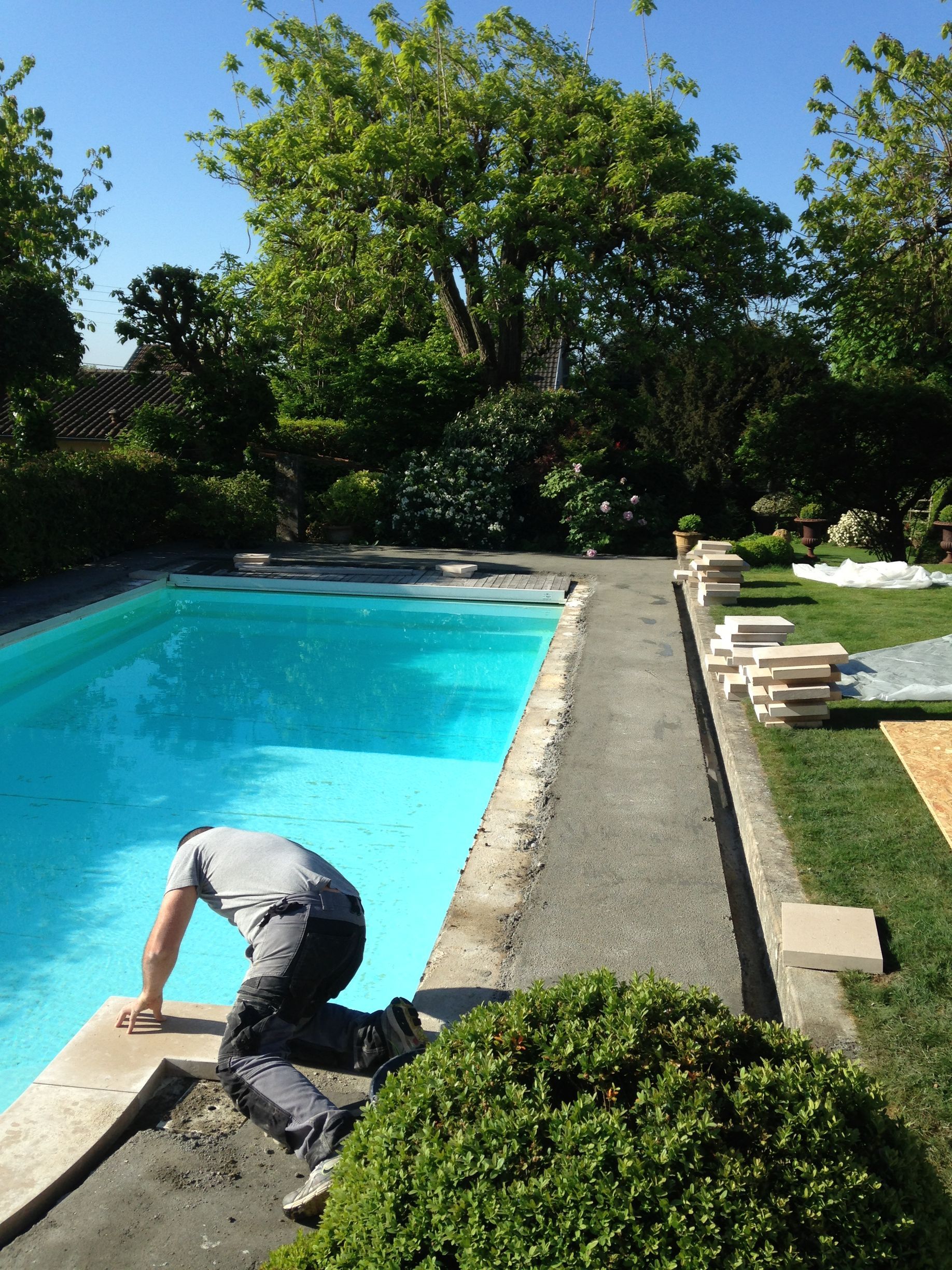Une personne accroupie au bord d'une piscine turquoise, à côté d'un chemin en béton et d'une pelouse verte dans un jardin ensoleillé.