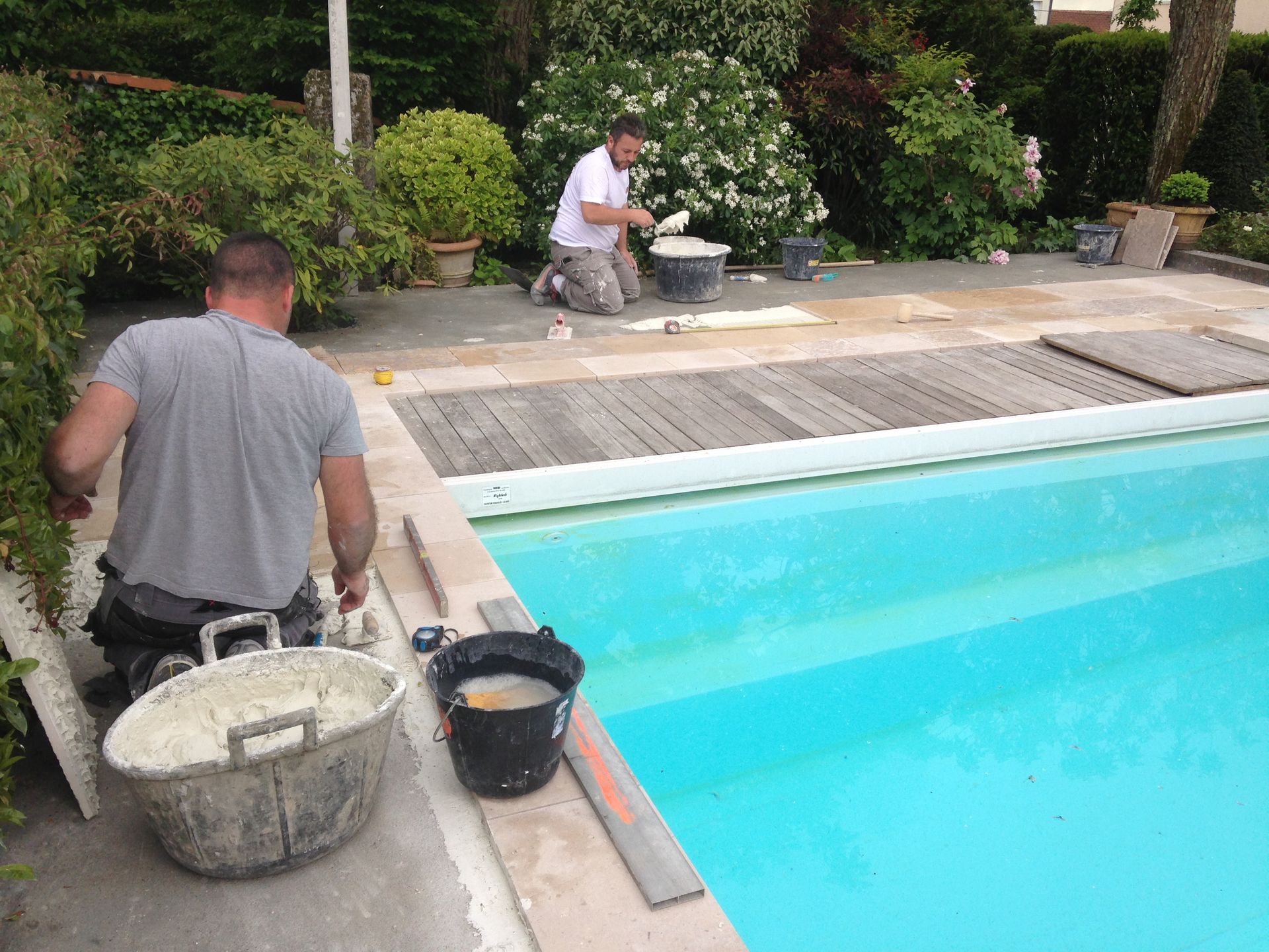 Deux ouvriers réparent la terrasse d'une piscine turquoise, avec des outils et des seaux à proximité.