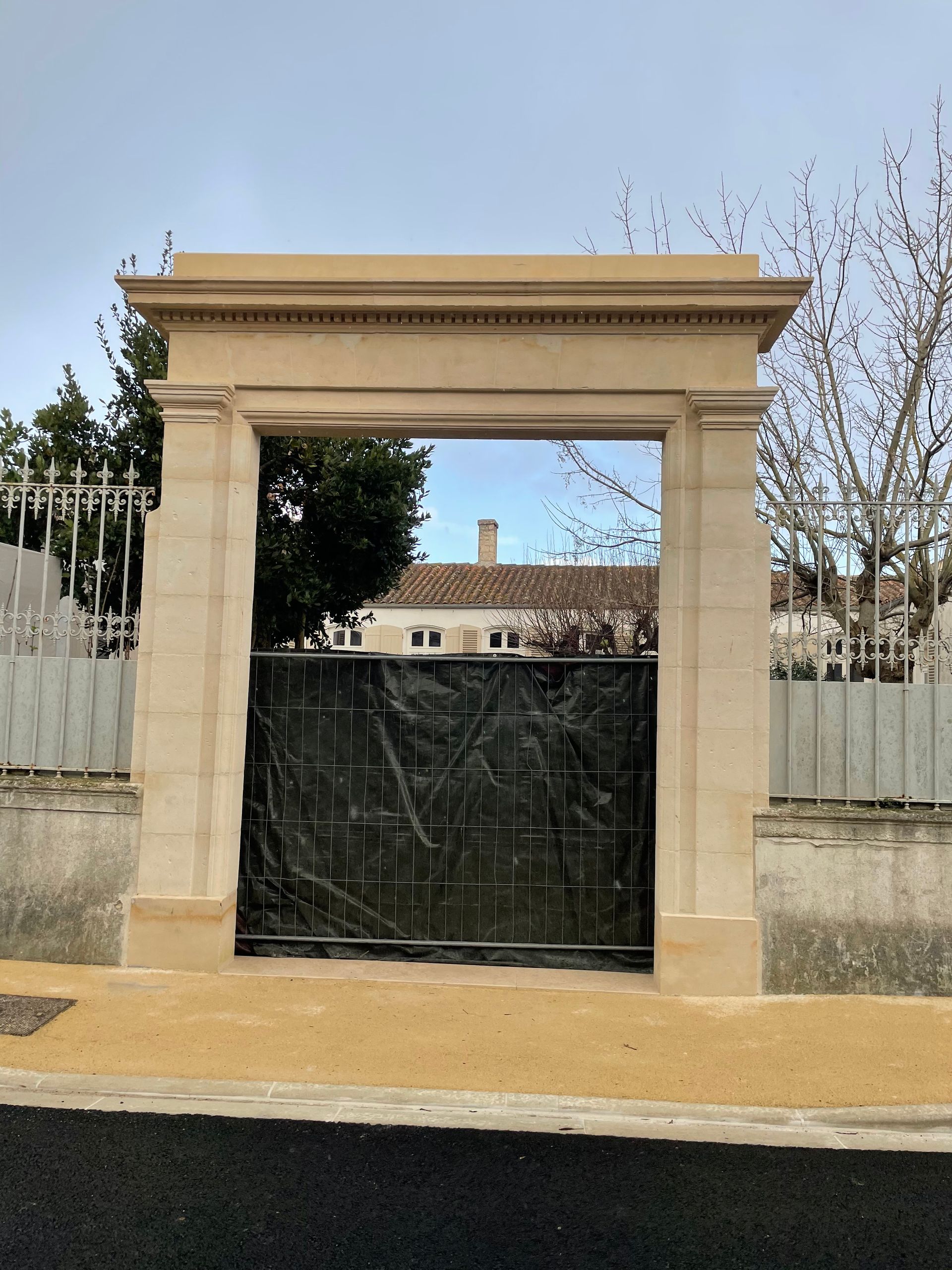 Portail d'entrée en pierre avec une grille en métal noir et des arbres en arrière-plan sous un ciel dégagé