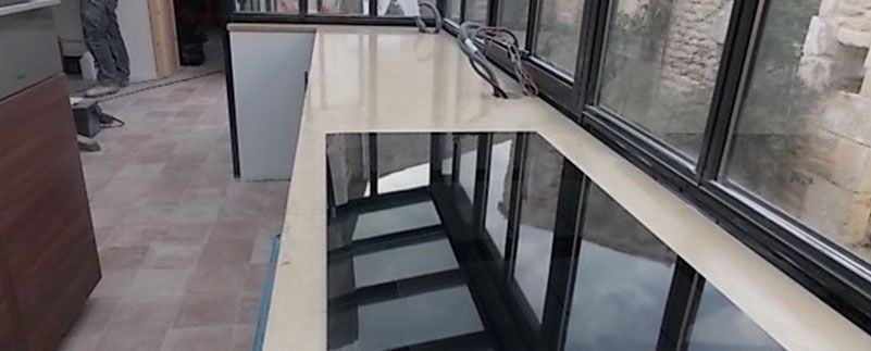 Îlot de cuisine moderne avec surface noire réfléchissante, à côté d'une grande fenêtre donnant sur un mur en pierre.