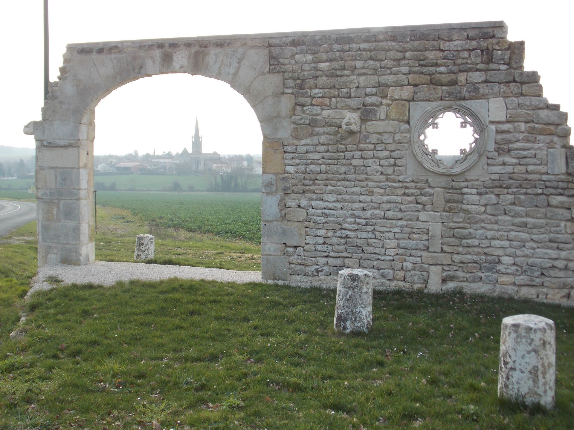 Ruine d'un mur de pierre avec une ouverture cintrée et une fenêtre circulaire donnant sur un champ herbeux et une ville au loin