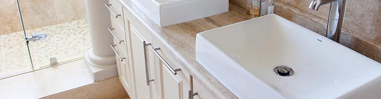 Salle de bains avec lavabo, robinet et meuble blancs. Mur carrelé de brun et sol en pierre.