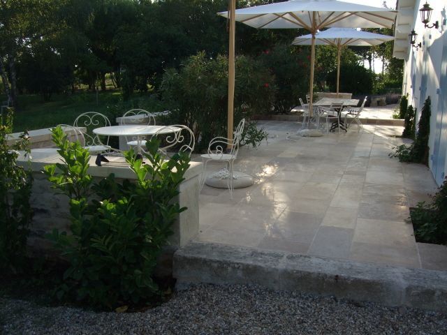 Terrasse avec tables et chaises blanches sous des parasols, à côté d'un bâtiment et de verdure.