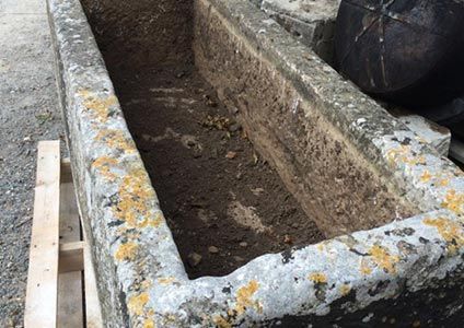 Abreuvoir rectangulaire en pierre, patiné par le temps, rempli de terre, posé sur une palette en bois.