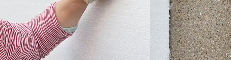 Le bras d'une personne vêtue d'une chemise rouge à rayures applique du mastic blanc sur un coin de mur.