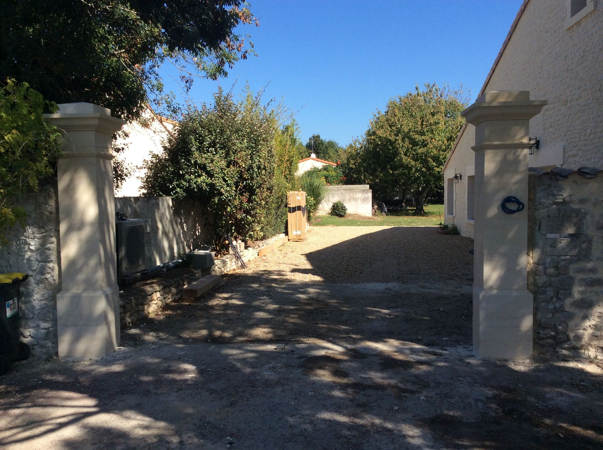 Allée pavée ensoleillée entre des murs et des piliers en stuc blanc, avec des arbres et une maison en arrière-plan