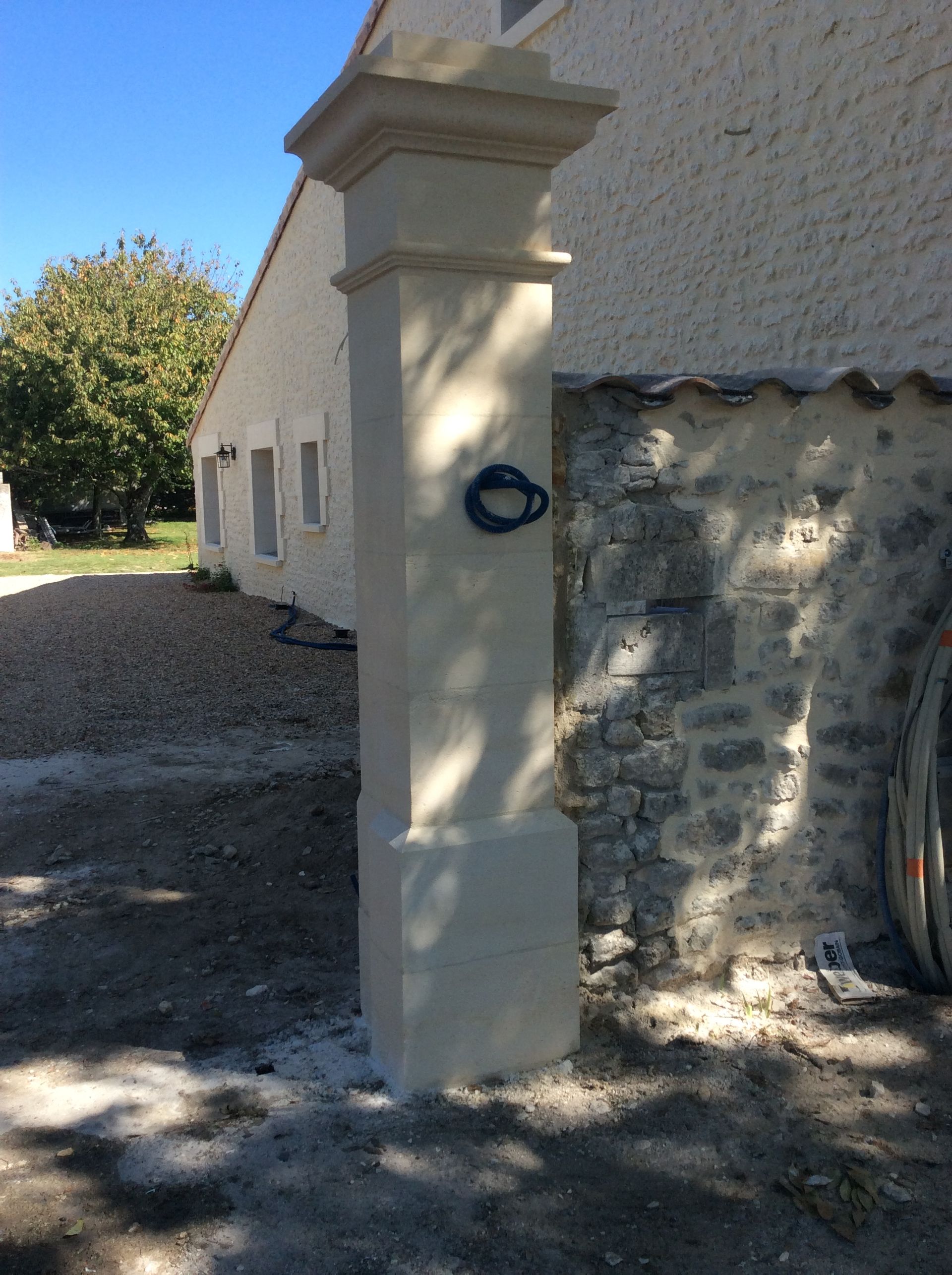 Pilier de pierre à côté d'un mur et d'un portail en stuc, avec une allée de gravier et des arbres baignés de soleil.