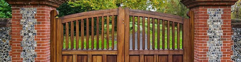Portail en bois entre des piliers de briques, avec un fond de feuillage vert.