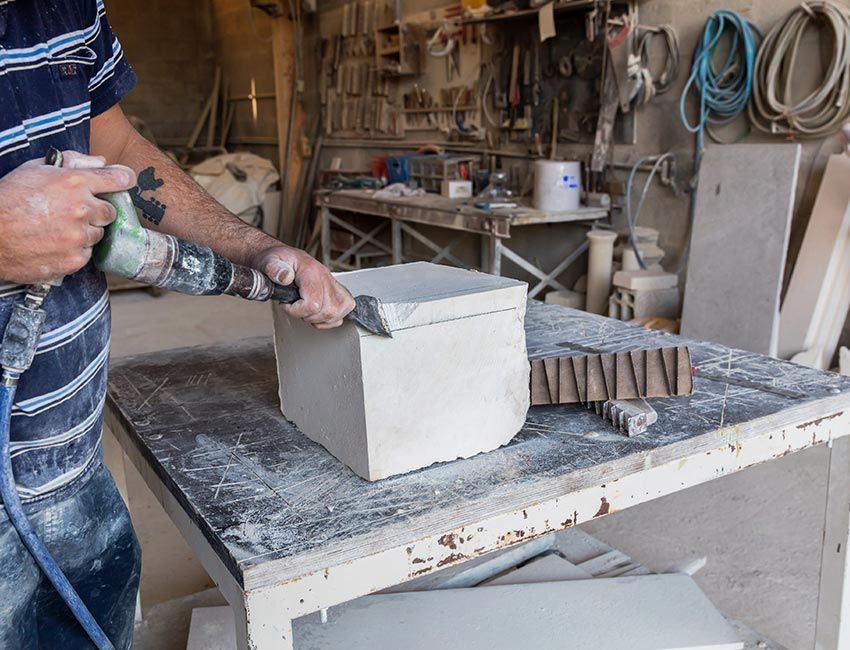 Une personne taille un bloc de pierre avec un outil dans un atelier. Des outils poussiéreux sont visibles en arrière-plan.