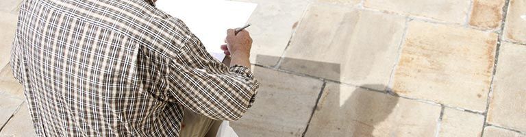 Une personne écrit sur du papier, près d'un mur de pierre. Elle porte une chemise à carreaux.