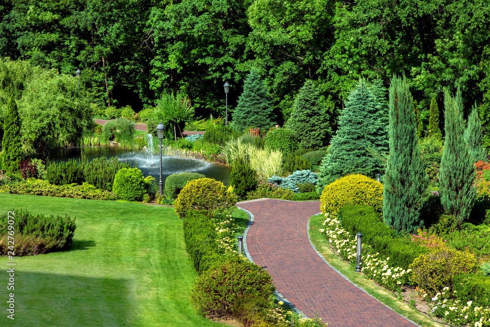 Un jardin soigné avec un chemin sinueux de gravier rouge, une pelouse verdoyante luxuriante, des arbustes taillés, de grands pins et une petite fontaine.