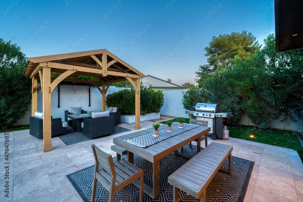 Une terrasse extérieure ensoleillée comprend un kiosque en bois avec des sièges de détente, une table à manger avec un banc et un barbecue en acier inoxydable.
