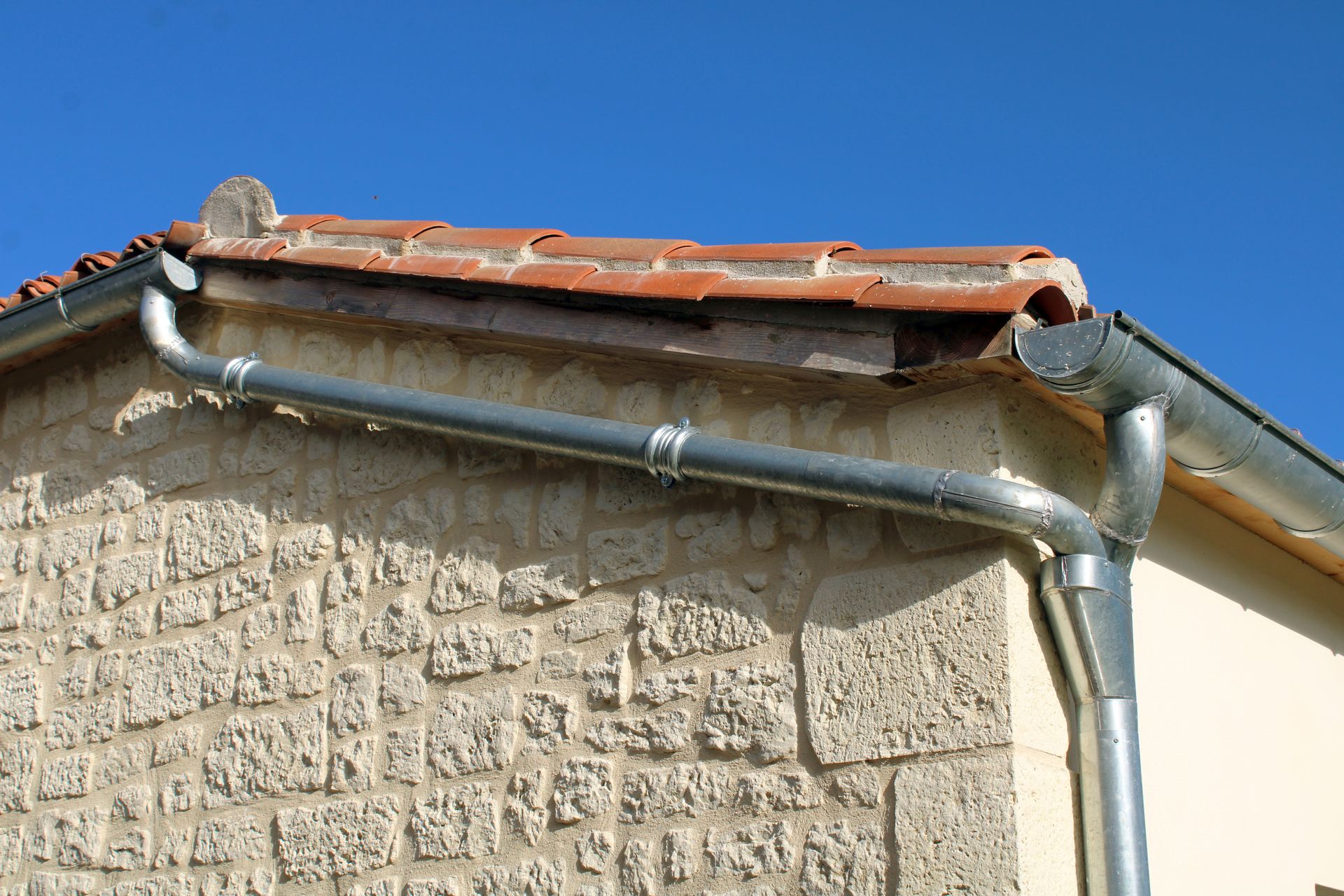 Gouttière en métal gris sur un bâtiment en briques au toit de tuiles en terre cuite, sous un ciel bleu dégagé.