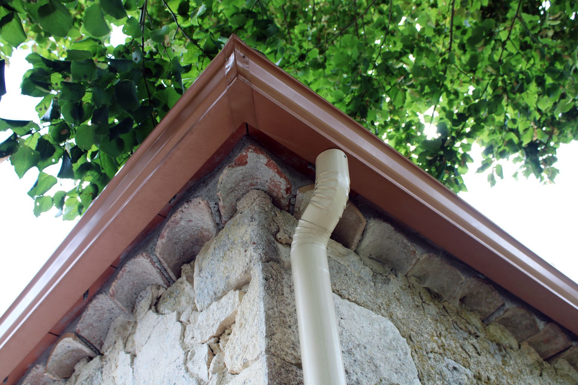 Gouttière et descente de toit brunes sur un bâtiment en pierre, avec du feuillage vert en arrière-plan.