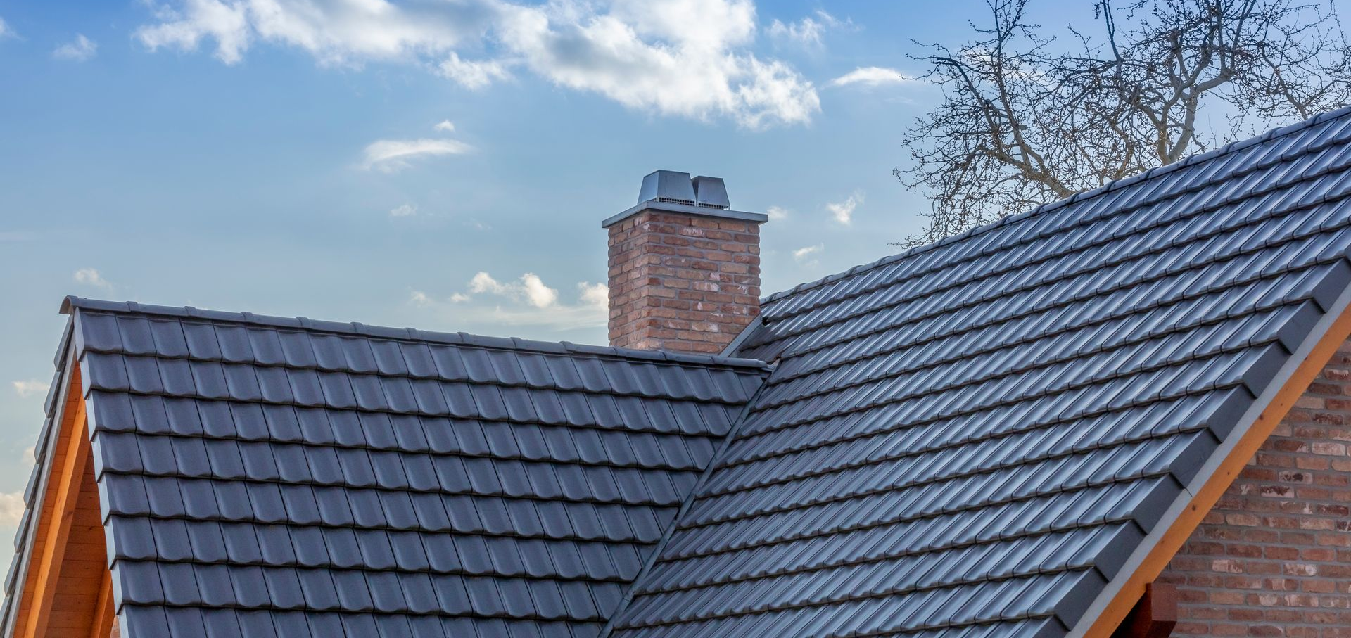 Toit de tuiles grises avec une cheminée en briques sur un ciel nuageux.